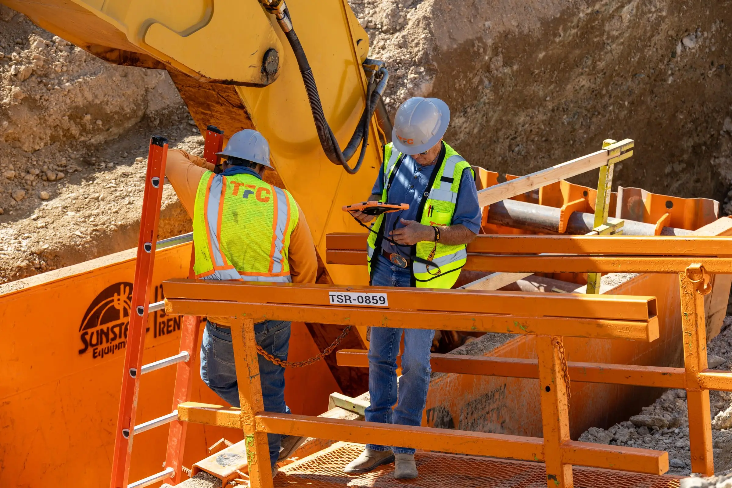 Two workers in hard hats reviewing plans inside a trench box