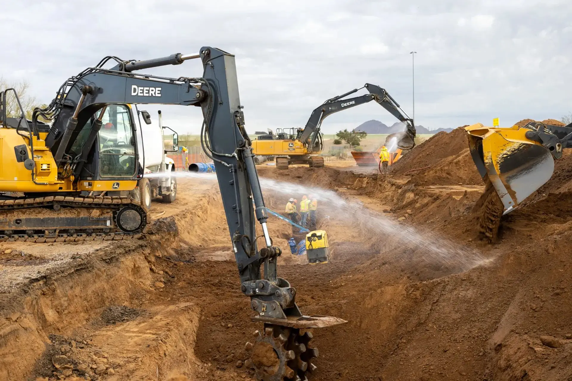 Excavator digging while a worker sprays water for dust control